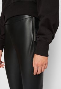 Zwarte leren broek met een gladde textuur, voorzien van een zijrits detail. De stof heeft een glanzende afwerking, gecombineerd met een donkere trui.