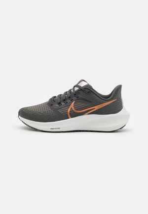 Grijze sportschoenen met een gestructureerde bovenkant, oranje swoosh-logo, witte tussenzool en zwarte rubberen buitenzool. Kenmerken zijn onder andere veters en een subtiel patroon.