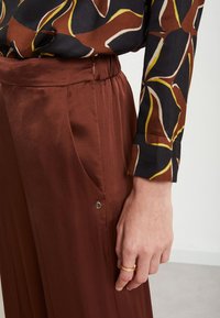 Pantalons en satin marron taille haute, dotés de poches latérales et d'un subtil détail métallique. Associés à un chemisier à manches longues à motifs.