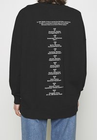 Sudadera negra con texto blanco en la parte posterior, detallando ubicaciones y fechas, con un corte relajado y puños acanalados.