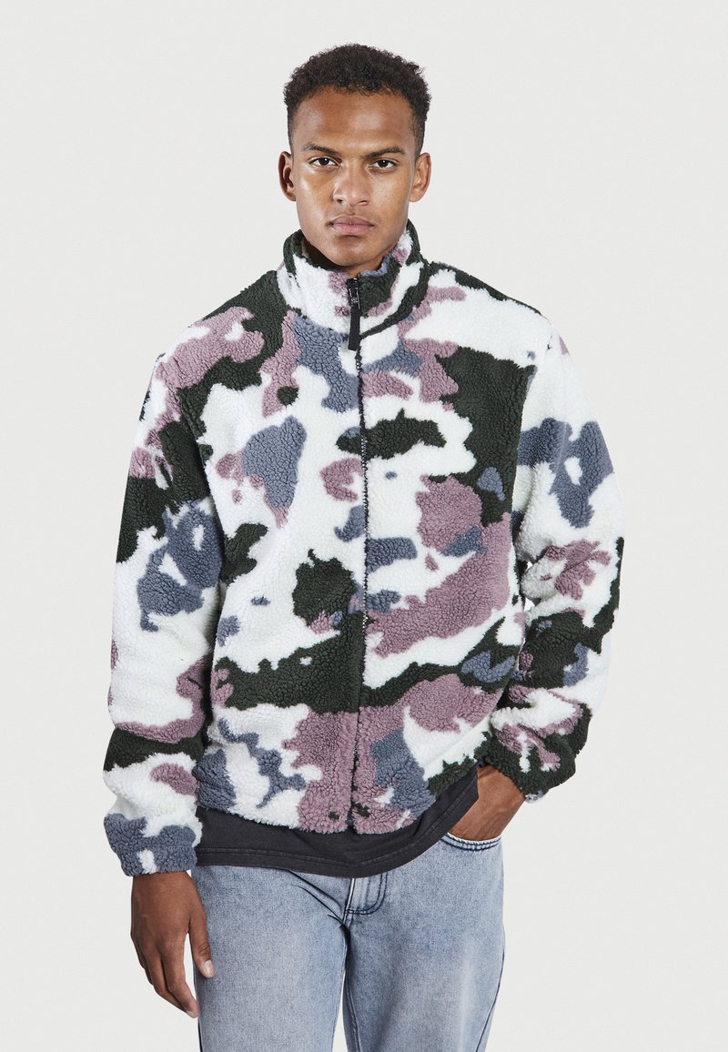 Mann trägt eine Fleecejacke mit Reißverschluss in einem mehrfarbigen Camouflage-Muster in Weiß, Grün, Grau und Lila, kombiniert mit hellblauen Jeans.