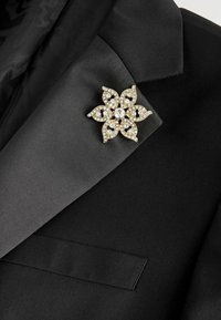 Goldene Blumenbrosche mit klaren Strasssteinen an einem schwarzen Smoking-Revers mit Satindetails und darunter liegender Brusttasche befestigt.