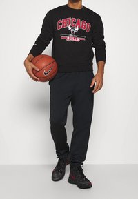 Felpa nera con grafica "Chicago Bulls", pantaloni della tuta neri e sneakers nere con dettagli rossi. Tiene una pallacanestro.