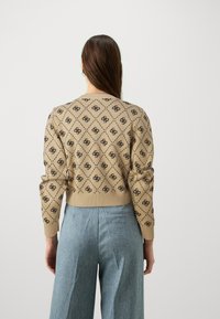 Jersey de punto beige con patrón de logo negro repetido, con puños y bajo acanalados, combinado con pantalones de pierna ancha azul claro.