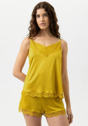 Junge Frau mit langen lockigen Haaren, die ein senfgelbes, mit Spitze verziertes Top und passende Shorts trägt, vor einem einfarbigen Hintergrund stehend.