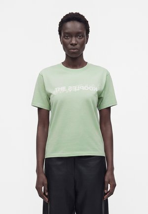 Kvinde med kort krøllet hår iført en lysegrøn T-shirt med hvidt forvrænget tekst og sorte bukser, stående mod en hvid baggrund.