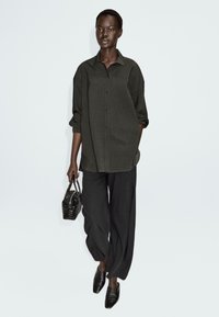 Camicia button-up oversize di colore verde scuro con maniche lunghe, abbinata a pantaloni neri larghi. Il modello tiene una piccola borsa in pelle intrecciata nera e indossa scarpe nere con punta.