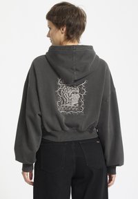 Grijze cropped hoodie met een groot, uitgebreid grafisch ontwerp op de achterkant. Heeft een capuchon en een strak gebreide tailleband. Gemaakt van zachte stof.
