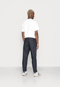 Camiseta branca de manga curta combinada com calças de ganga azul-escuro. O modelo está de lado, mostrando um ajuste descontraído e calçado desportivo casual.