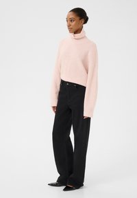 Roza pletena turtleneck pulover z ohlapnimi rokavi, v kombinaciji z visokim pasom črnimi kavbojkami in črnimi špicastimi čevlji. Gladki, teksturirani materiali.