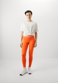 T-shirt noué blanc, leggings ajustés orange vif avec logo Nike blanc, et chaussures de sport blanches. Design simple et texture lisse.