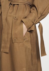 Brązowa kurtka z paskiem o gładkiej fakturze, z dwoma dużymi guzikami i bocznymi kieszeniami, zaprojektowana z czystą i dopasowaną sylwetką.