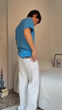 Fiatal férfi kék pólóban és fehér nadrágban, egy minimalista hálószobában áll az ágy mellett, közel egy keretezett képhez és gyertyatartóhoz.