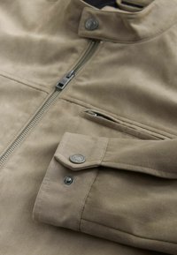 Giacca in camoscio beige con chiusura a zip, bottoni a pressione su colletto e polsino, e una tasca frontale. Tessuto morbido e superficie liscia.