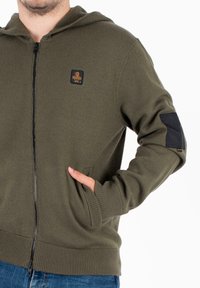 Felpa con zip di colore verde oliva, realizzata in tessuto morbido, con tasca frontale, polsini a coste e una toppa nera sulla manica sinistra.