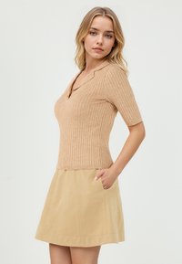 Pull en tricot côtelé beige à manches courtes, avec un col en V et un design ajusté, associé à une jupe évasée beige avec des poches latérales.