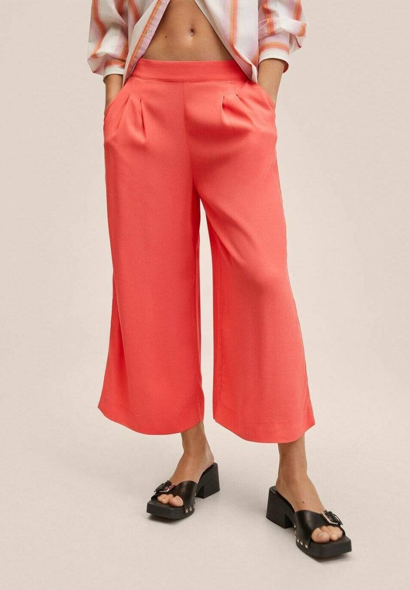 Koraalrode wijde broek met een zachte textuur, voorzien van zijzakken en plooien. Gecombineerd met zwarte sandalen met blokhakken.