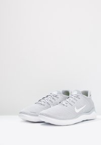 Jasnoszare buty sportowe z teksturowaną siateczką u góry, sznurowane, z białą podeszwą, na której widoczny jest wyraźny biały logo.