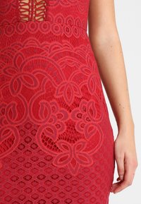 Rotes Spitzenkleid mit aufwendigen Blumenmustern, taillierter Passform und strukturierter Oberfläche. Ein Ausschnitt am Kragen betont das Gesamtbild.