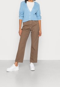 Ljusblå stickad cardigan med V-hals, knappar, i kombination med bruna rakbenade byxor och vita sneakers mot en enfärgad bakgrund.