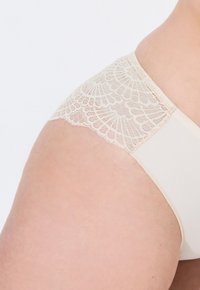 Beige spodní prádlo s květinovým krajkovým lemem na pase, s hladkým povrchem a bezešvým designem sahajícím k bokům.