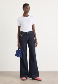Femme portant un haut blanc à manches courtes, un jean large foncé, des baskets roses, tenant un petit sac à main bleu, debout sur un fond clair uni.
