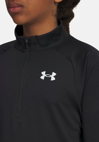 Person iført en sort zip-up sportsbluse med et hvidt Under Armour-logo på venstre bryst, vist fra halsen til midten af torso.