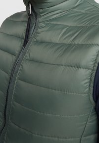 Vadderad väst i mörkgrön nylon med kviltade sektioner, hög krage och dragkedja fram, med en slät, glänsande yta.
