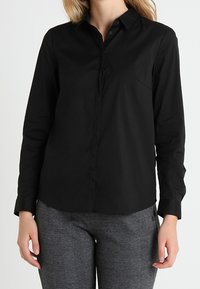 Chemise noire à manches longues avec un col boutonné, une poche poitrine unique et une texture de tissu lisse. Associée à un pantalon gris à carreaux.