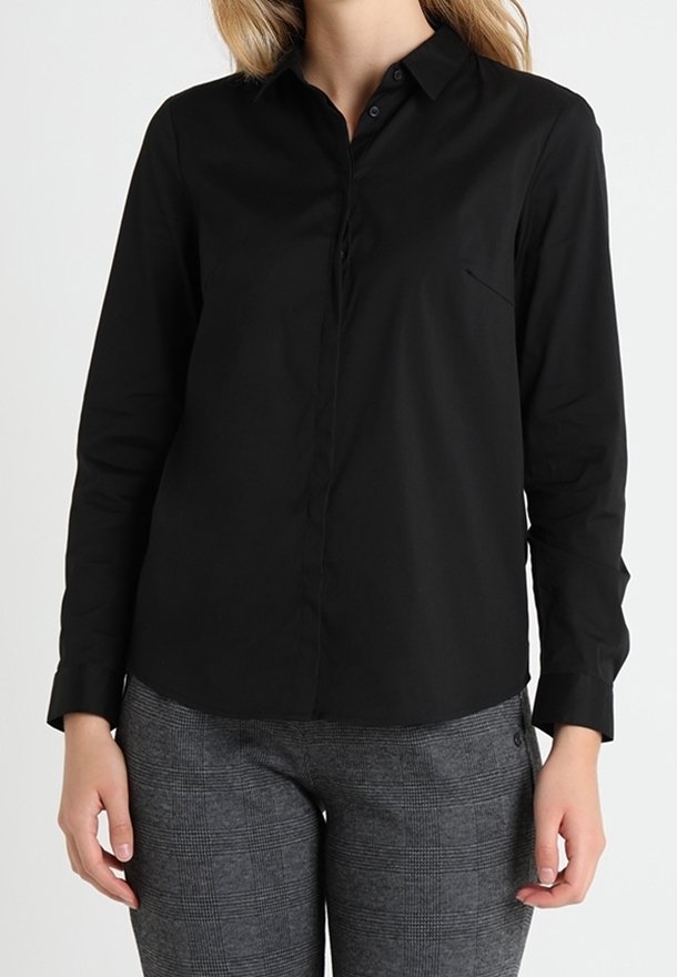 Chemise noire à manches longues avec un col boutonné, une poche poitrine unique et une texture de tissu lisse. Associée à un pantalon gris à carreaux.