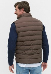 Brązowa kamizelka puchowa z poziomym pikowaniem, wysoki kołnierz, noszona na niebieskiej koszuli z długim rękawem, widok z tyłu. Luźny krój i design.