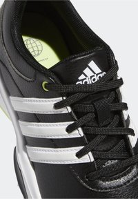 Czarne buty sportowe z białymi paskami, górna część z teksturowanej syntetycznej skóry, czarne sznurowadła i zielona wkładka z logo.