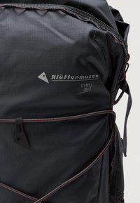 Črna nahrbtnik s kapaciteto 35L, ki ima teksturirano površino, subtilno sivo blagovno znamko, šivano rdeče poudarke in zasnovo z zategnjenim zapiranjem.