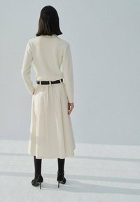 Cream, fitted sweater with long sleeves paired with a floor-length, pleated, cream skirt. Black belt at the waist, black tights, and high-heeled shoes.