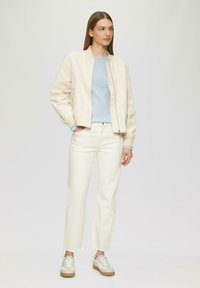 Veste bomber beige clair sur un t-shirt bleu clair, accompagnée de jeans droits blancs et de baskets crème avec une semelle marron.