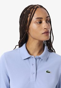 Polo shirt azzurro chiaro in cotone, con colletto classico e tre bottoni. Nota il logo verde del coccodrillo sul petto.