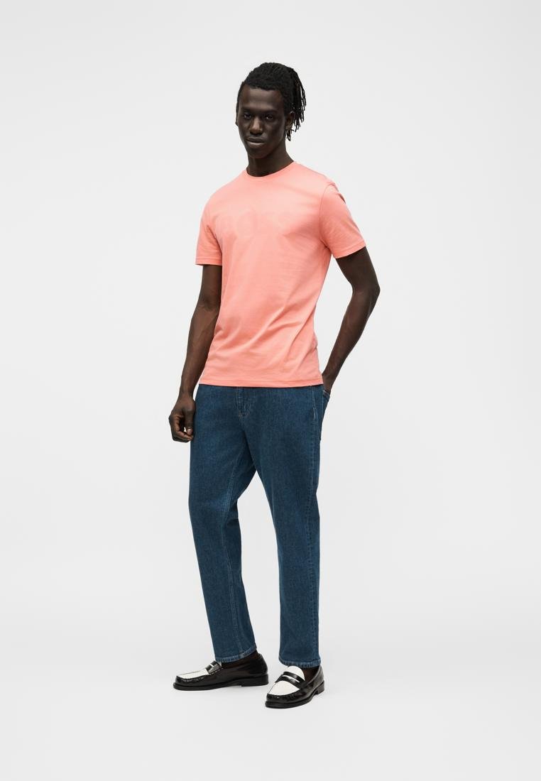 Homme à la peau foncée et aux dreadlocks portant un t-shirt corail à manches courtes, un jean bleu et des mocassins noir et blanc, debout sur un fond uni.