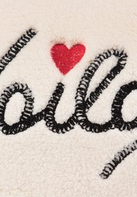 Tela de crema suave con texto en cursiva bordado en negro que dice "Holly" con un pequeño acento de corazón rojo. Superficie texturizada y esponjosa.