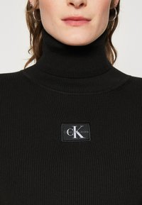 Maglione nero a coste con colletto alto, caratterizzato da un patch quadrato in bianco e grigio. Tessuto liscio, design aderente.