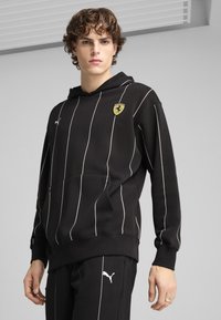 Crna dukserica s kapuljačom s bijelim vertikalnim prugama, koja ima prednji džep i Ferrari logo na prsima. Mekani, teksturirani materijal.