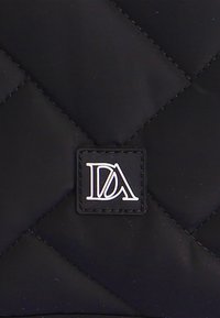 Tejido negro acolchado con un patrón de diamantes, con un pequeño parche de cuero negro con letras blancas en relieve "DA" en una fuente estilizada.