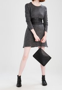 Abito lungo grigio a maniche lunghe con una cintura in vita texturizzata. Il modello tiene una pochette geometrica nera in finta pelle, abbinata a stivali neri alla caviglia.