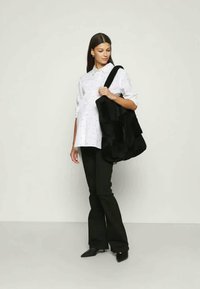 Saco de tecido preto com design em camadas. Modelo veste uma camisa branca de botão com bolinhas pretas e calças pretas largas. Textura suave.