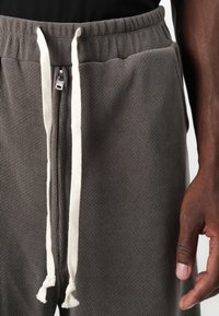 Gray waffle-textured sweatpants with an elastic waistband, featuring a white drawstring and metal zipper detail. Close-up view of waist area.