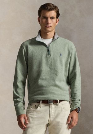 Homme portant un pull vert clair à fermeture éclair sur une chemise blanche, pantalon beige, ceinture marron et montre au poignet gauche, debout devant un fond uni.