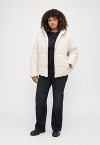 Ανοιχτό μπεζ μπουφάν puffer με κουκούλα, με ένα επενδυμένο σχέδιο, μπροστινές τσέπες και φερμουάρ. Φορεμένο πάνω από μαύρη μπλούζα και μαύρο παντελόνι.