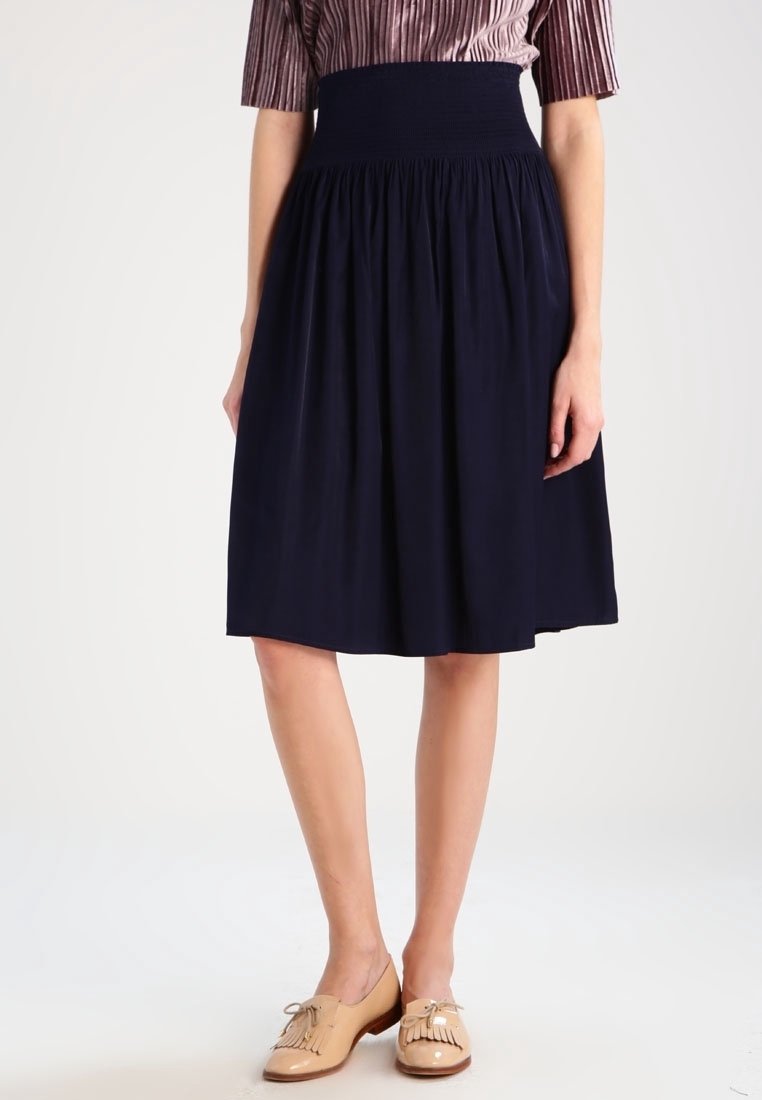 Woman wearing a high-waisted, navy blue midi skirt paired with beige tassel loafers, standing against a plain white background.