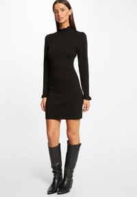 Vestido negro de punto con mangas largas, un cuello alto con volantes y una silueta ajustada. Lleva zapatos de cuero negro hasta la rodilla.