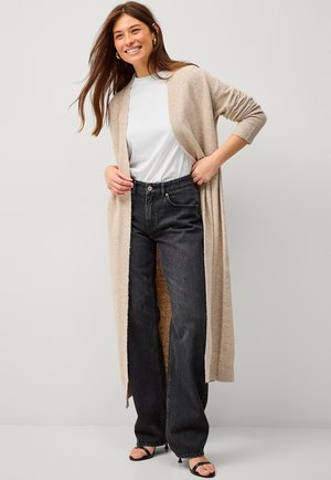 Beige, lange, open cardigan van zachte stof over een wit t-shirt, gecombineerd met hoge zwarte jeans en zwarte sandalen.