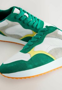 Adidași verzi și albi cu panouri din suede și piele. Prezintă accente galbene și o talpă din cauciuc texturată cu branhii portocalii. Design athletic.